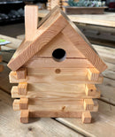 A wooden bird box designed to resemble a small log cabin with a chimney stack and a gable roof.