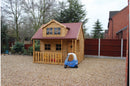 Children's wooden mansion playhouse with a veranda, dormer window, and a blue toy car in front, set on a gravel surface in a garden.