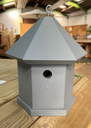A gray hexagonal bird box designed to resemble a house with a entrance hole on the front.
